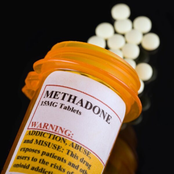 Bottle of Methadone oral solution with dosage label, alongside a plastic dose cup on a pharmacy counter.